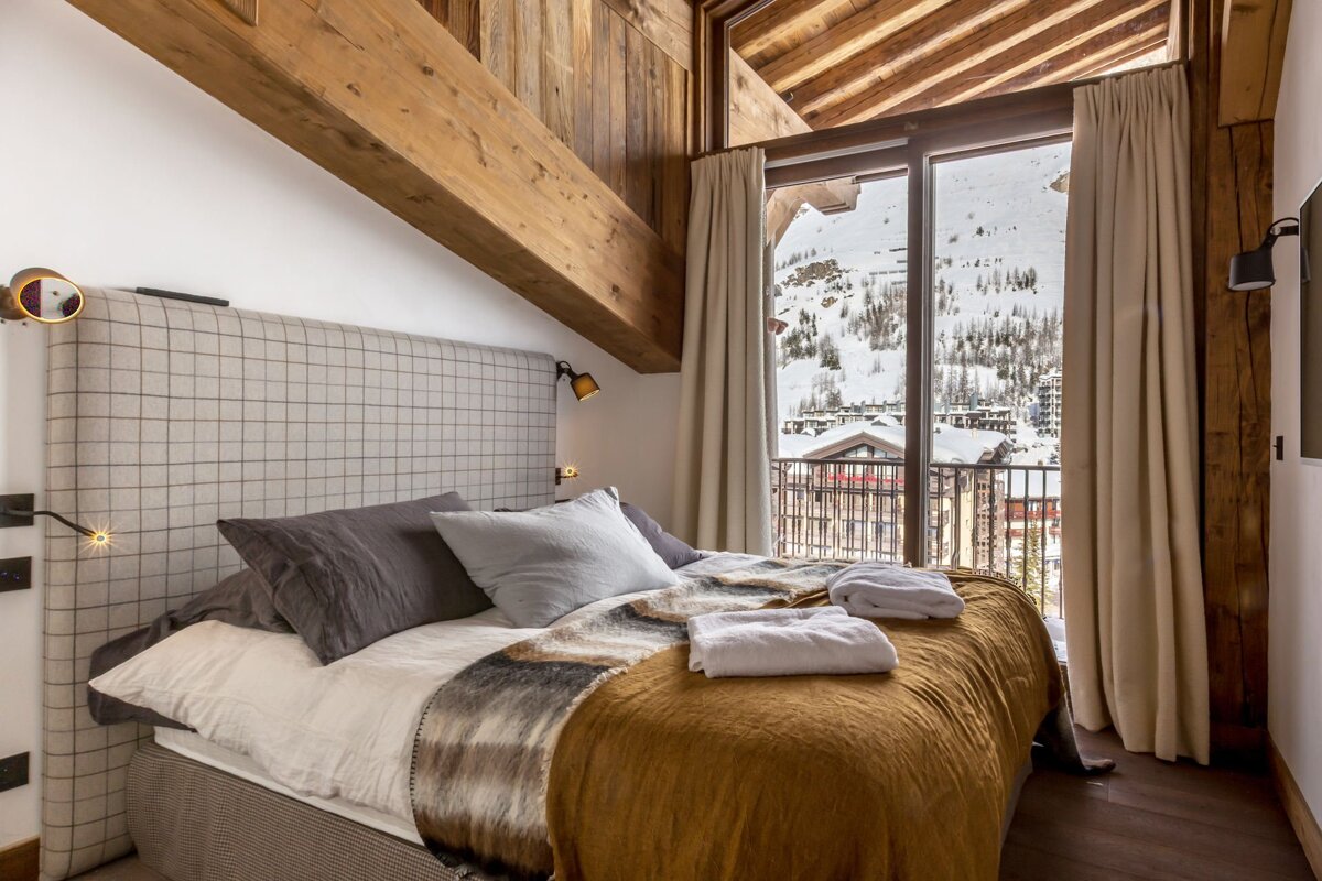 Cozy chalet bedroom featuring wooden beams, a plush bed, and large windows overlooking a snowy mountain village.