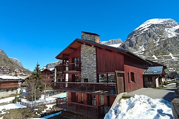 A multi-story wooden chalet with stone chimney and balconies, nestled in a snowy mountain village under a bright blue sky.