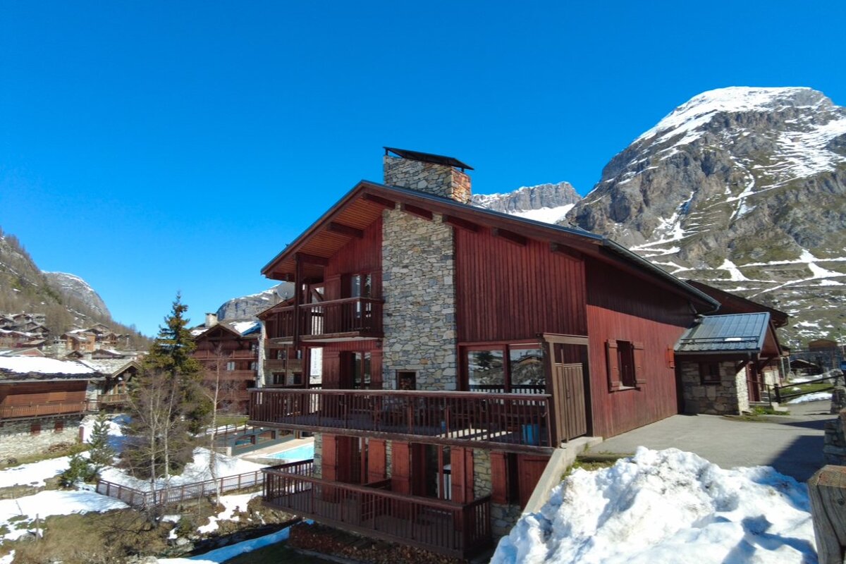 A wooden and stone chalet in a snowy alpine landscape with towering mountains under a bright blue sky.