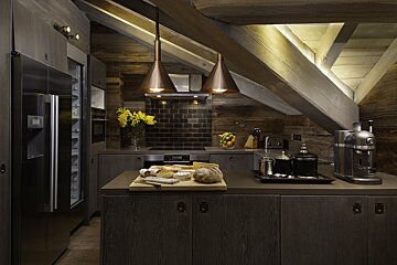 A modern rustic kitchen with dark wood, exposed beams, and sleek appliances. Warm lighting highlights the angled ceiling and a breakfast spread.