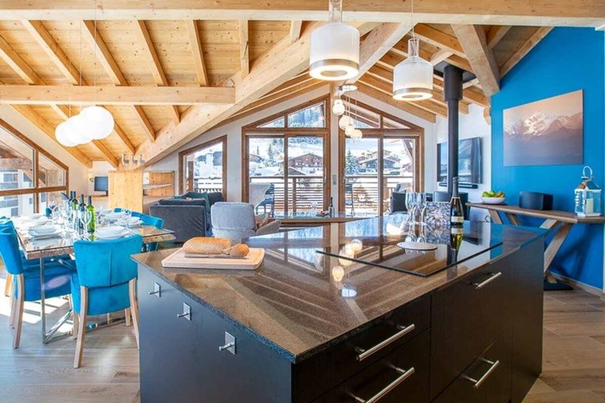 Modern chalet interior with vaulted wooden ceilings, vibrant blue chairs, a sleek island, and windows overlooking a snowy mountain resort.