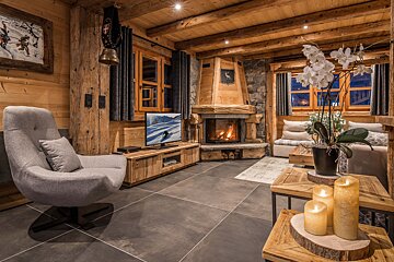 Cozy rustic chalet living room with a stone fireplace, wood-paneled walls, a grey armchair, and candles on wooden tables.