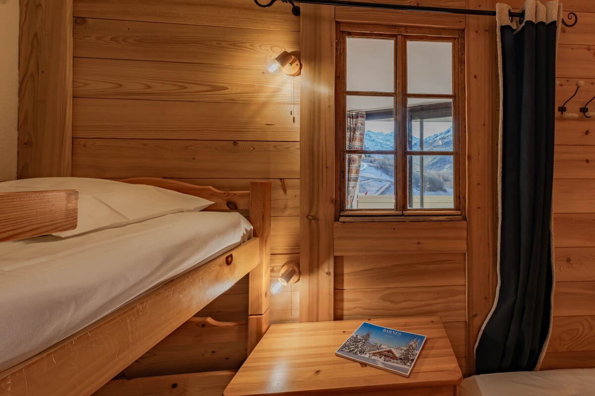 Cozy chalet bedroom with wooden bunk beds, warm lighting, and a window view of snowy mountains.