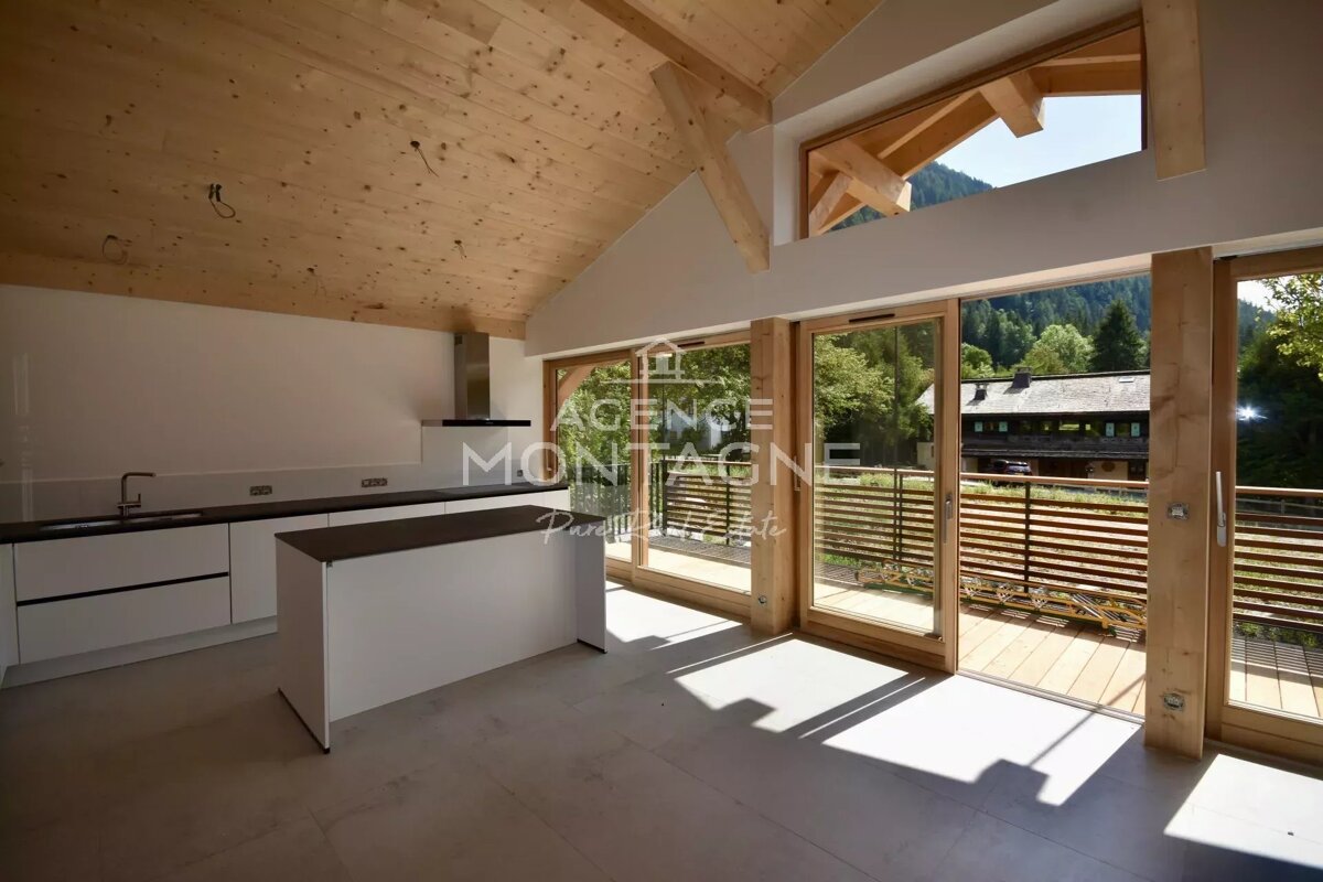 A bright, modern kitchen with a wooden vaulted ceiling and island. Large glass doors open to a balcony with scenic mountain and forest views.