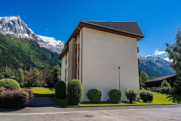 A white building with a brown roof stands against a stunning backdrop of snow-capped mountains under a bright blue sky.