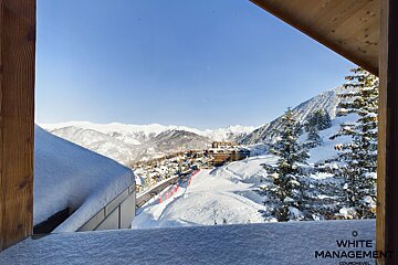 A stunning view of a snowy ski resort village nestled among majestic mountains and pine trees, seen from a window under a clear blue sky.