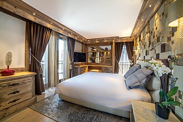 A stylish chalet-style bedroom with a large white bed, warm wooden accents, a patterned wall behind the bed, and large windows.