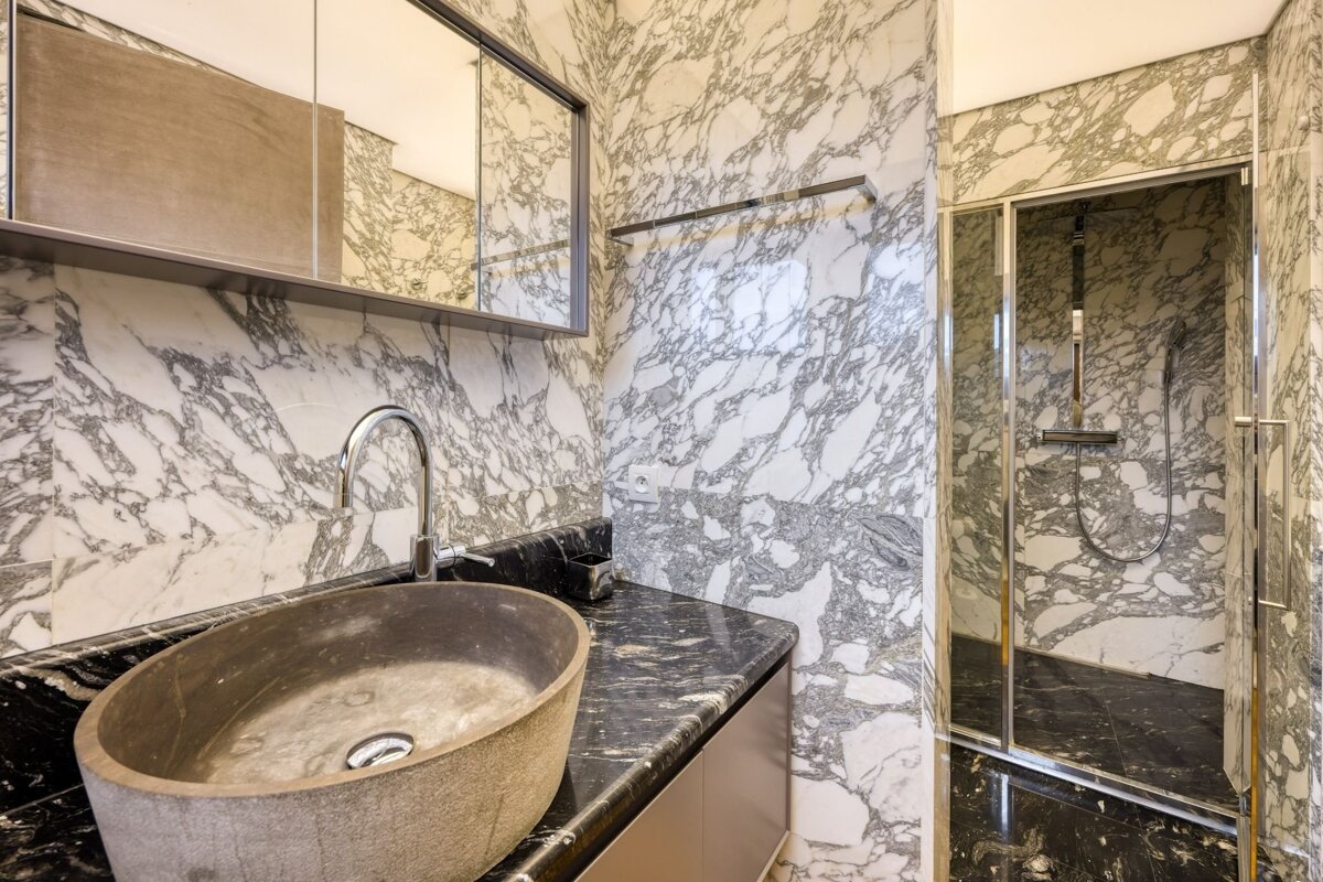 A luxurious marble bathroom featuring white-veined walls, a black marble counter with a stone vessel sink, and a glass shower.