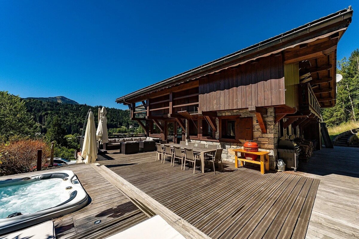 A spacious wooden chalet with a large deck, hot tub, and outdoor dining, overlooking a forested mountain range under a clear blue sky.