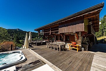A spacious wooden chalet with a large deck, hot tub, and outdoor dining, overlooking a forested mountain range under a clear blue sky.