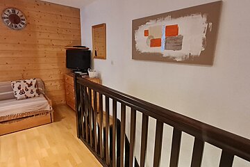A cozy room features a wood-paneled wall with a daybed and TV, opposite a white wall with abstract art, beside a dark wooden stair railing.