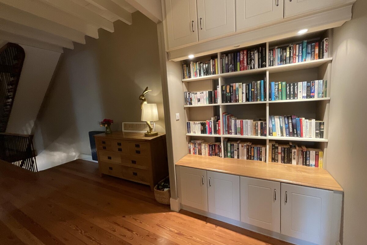 A cozy hallway with a large built-in bookshelf filled with books, a wooden dresser with a rabbit lamp, and warm wooden floors. A stairwell is visible nearby.