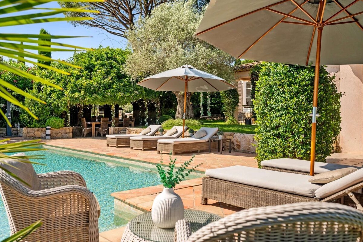 A luxurious poolside patio with sun loungers, umbrellas, and lush greenery creates a serene outdoor retreat under a sunny sky.