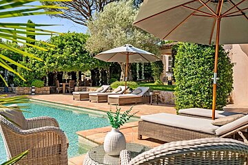 A luxurious poolside patio with sun loungers, umbrellas, and lush greenery creates a serene outdoor retreat under a sunny sky.