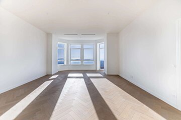 Bright, empty room with white walls and herringbone wood floor. A bay window shows mountains, and sunlight creates long shadows.