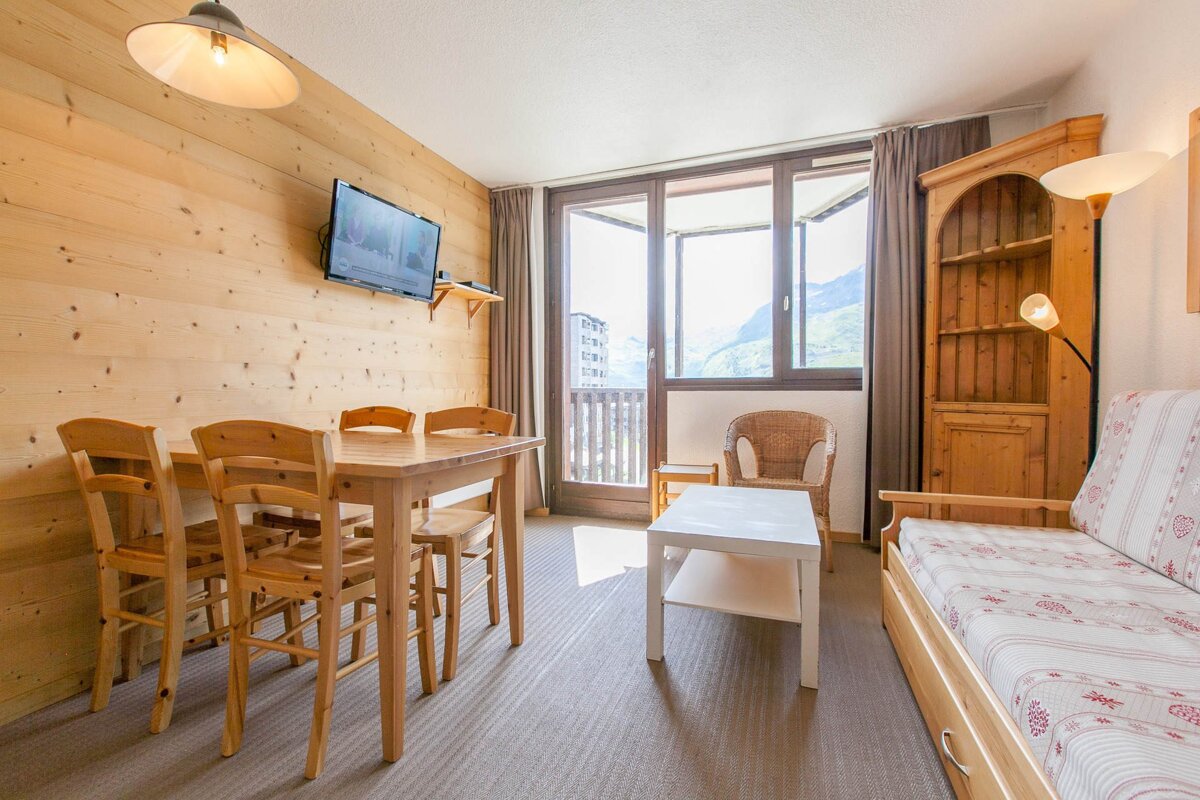 Cozy living/dining room with wood paneling, dining table, sofa bed, TV, and a large window/balcony overlooking mountains.