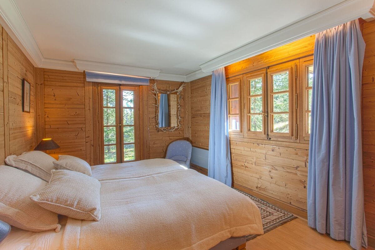A cozy, rustic wood-paneled bedroom with a large bed, blue curtains, and windows overlooking a forest. An antler-framed mirror hangs on the wall.