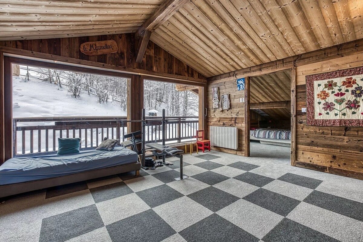 A cozy, wood-paneled cabin room with a checkered floor, a bed, a workout bench, and a snowy forest view through large windows.