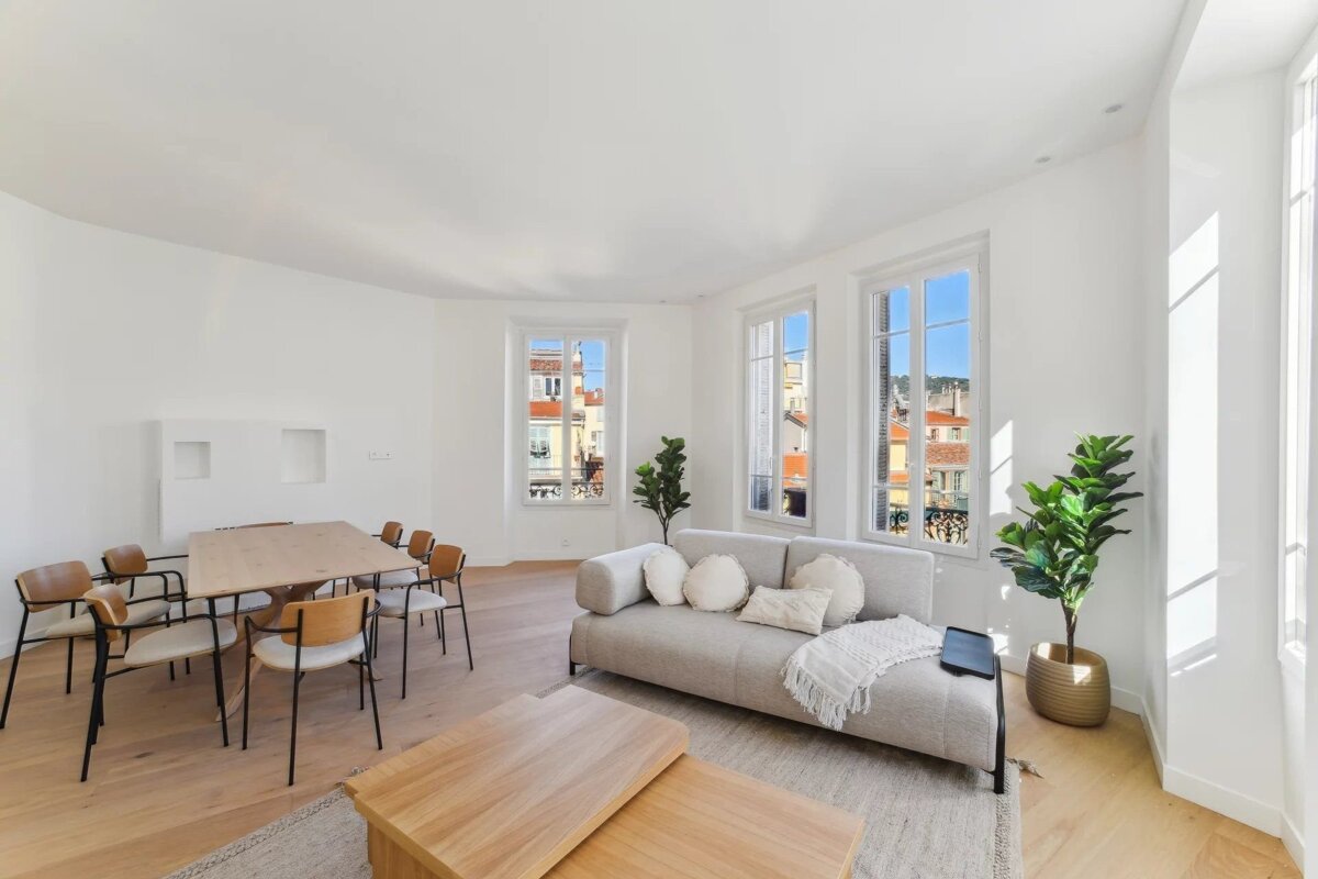 Bright, modern living and dining room with white walls, wood floors, a grey sofa, wooden table, and tall windows.