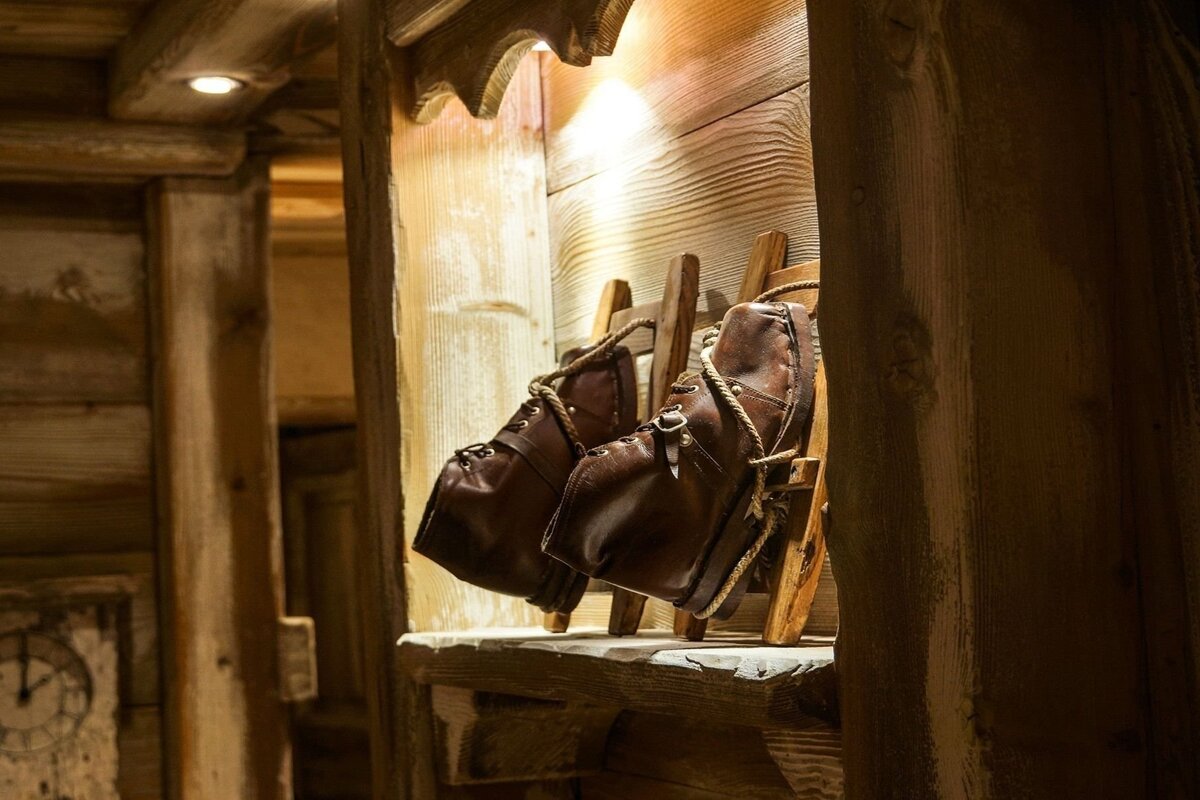 Old brown leather boots with rope laces sit on wooden stretchers within a rustic wooden display, lit by a warm light.