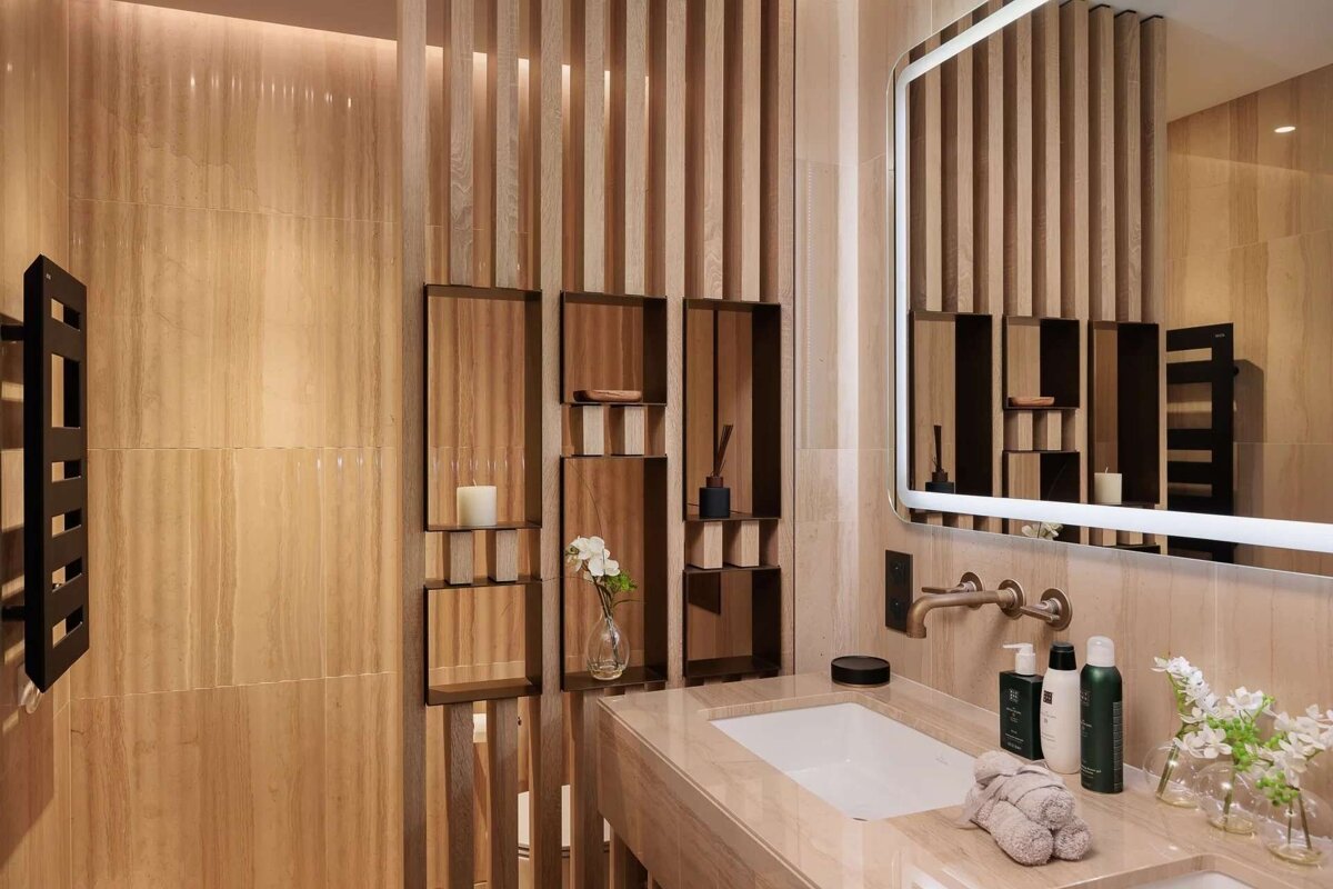 A modern bathroom featuring warm wooden slat screens, light marble walls, a sink, an illuminated mirror, and toiletries.