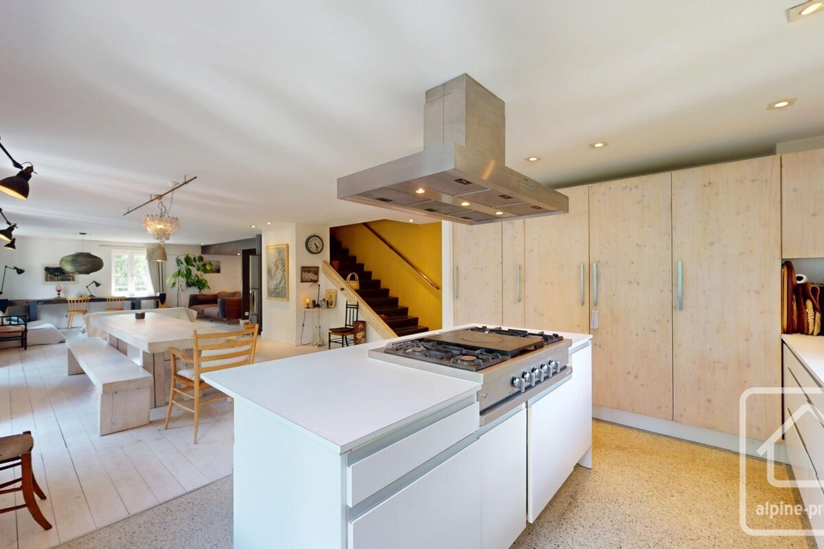 A modern open-concept living space featuring a white kitchen island with a gas range, light wood cabinetry, and a dining area. Yellow stairs lead up in the background.