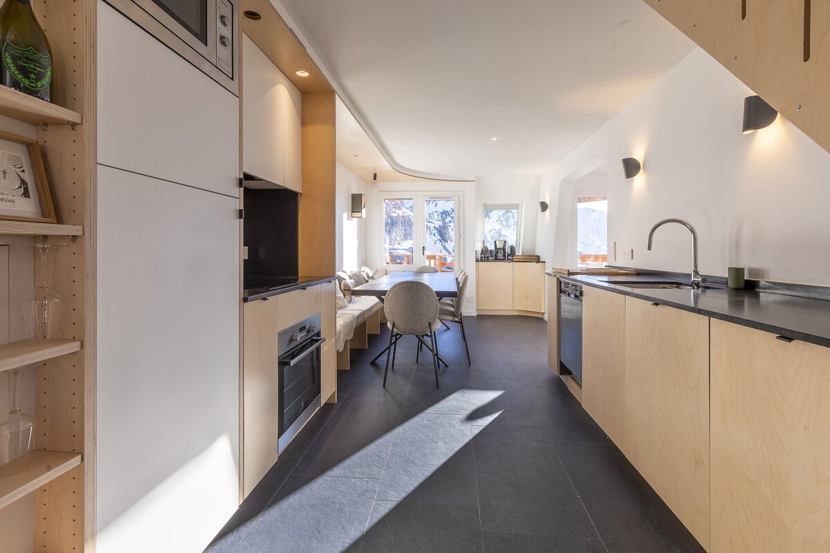 A bright, modern kitchen and dining area features light wood cabinetry, white appliances, a dark floor, and stunning snowy mountain views.