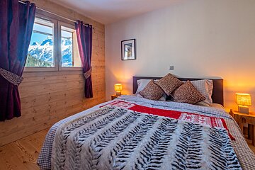 A cozy, rustic bedroom with wood walls, a comfy bed, warm lamps, and a window revealing a stunning snow-capped mountain view.
