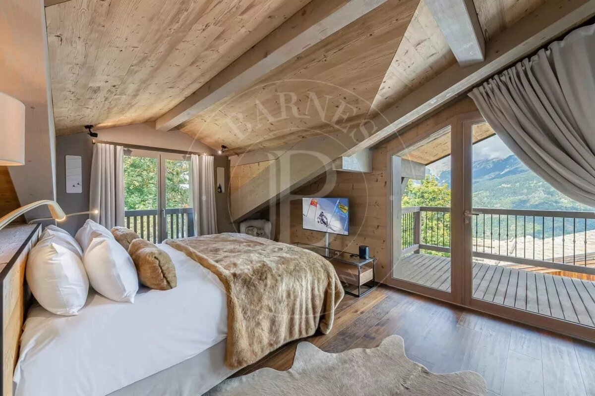 Cozy chalet bedroom with wood decor, a large bed, and two balconies offering panoramic mountain and forest views. A TV displays a skiing scene.
