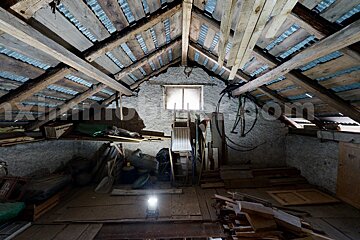 A rustic, cluttered attic with exposed wooden beams, a corrugated metal roof, and a window. Miscellaneous items and tools are stored, illuminated by a floor light.