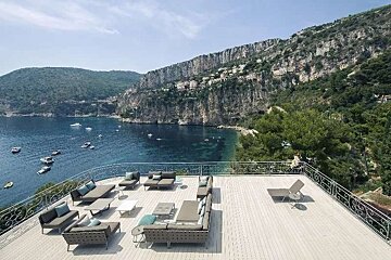 Luxurious outdoor terrace with modern lounge seating offers stunning views of a serene blue bay with boats, backed by dramatic cliffs and mountains.