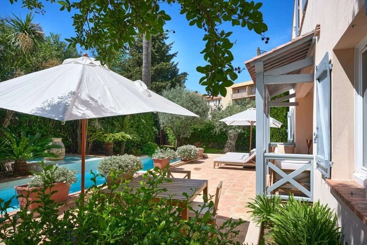 Sunny patio with a blue swimming pool, shaded by white umbrellas, next to a house with a porch, surrounded by lush green foliage.