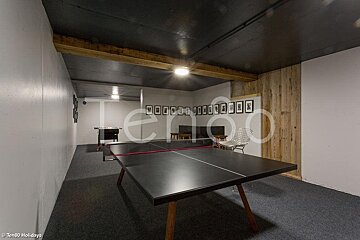 A recreation room with a black ping pong table, grey carpeting, wooden accents, and a foosball table in the background.