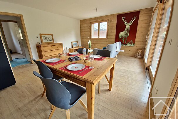 Cozy modern rustic dining/living room. Features a set wooden table, grey sofa, wood-paneled wall with deer art, and light wood floors.
