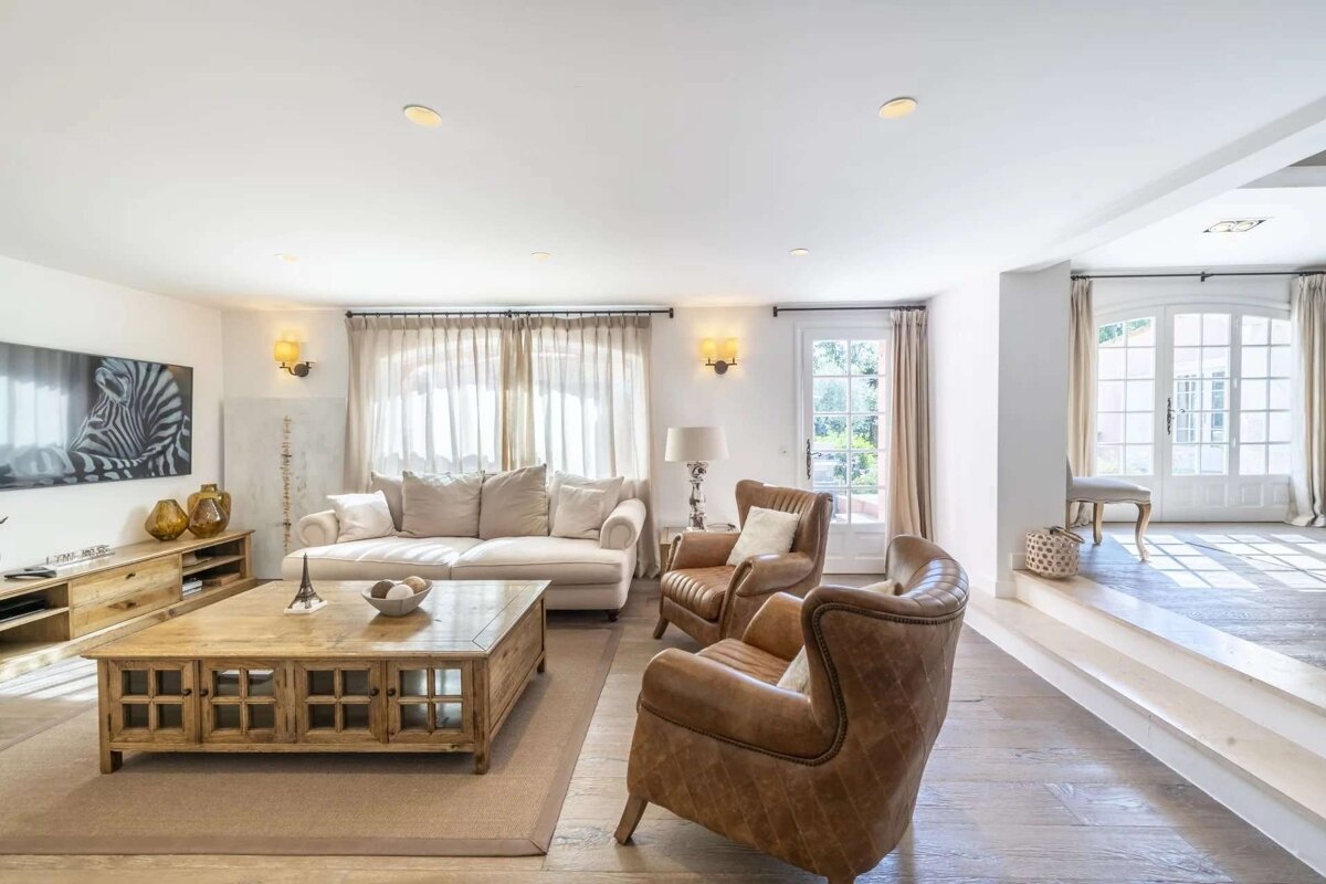 A bright living room with a cream sofa, brown leather armchairs, a wooden coffee table, and large French doors leading to another area.