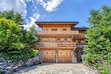 A rustic wooden chalet with stone base, multiple balconies, and two wooden garage doors, nestled among green trees under a blue, cloudy sky.