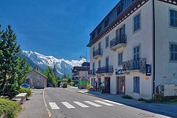 An alpine street with blue-shuttered buildings leading towards majestic, snow-capped mountains under a clear blue sky.