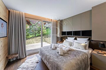 A modern, neutral-toned bedroom features a large bed with a faux fur throw, floor-to-ceiling glass doors opening to a green outdoor area, and wood paneling.