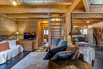 Cozy, rustic wooden living room and kitchen with sofas, TV, coffee table, an open staircase, dark tiled floors, and warm lighting.