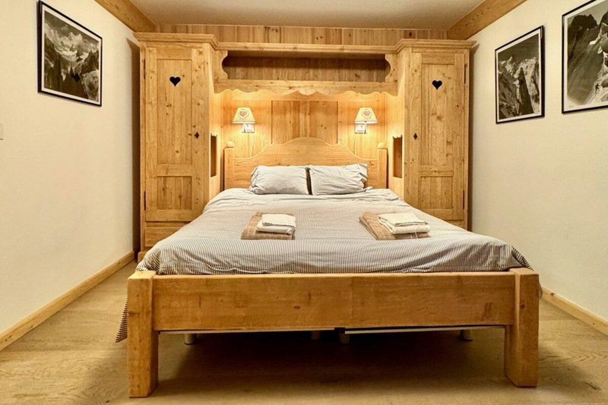 Cozy chalet-style bedroom with a light wooden bed built into matching wardrobes with heart cutouts and glowing wall lamps.