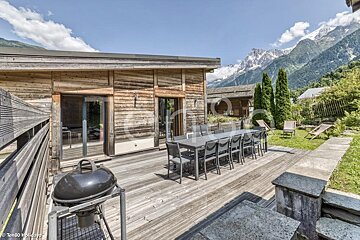 Wooden chalet deck with a large dining table and a BBQ grill, offering views of stunning, snow-capped mountains.