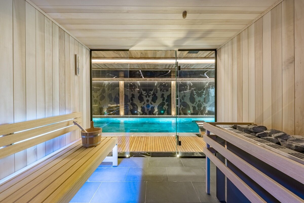 A modern sauna room with light wood walls, bench, and heater, offering a view through a glass partition to a blue-lit indoor swimming pool.