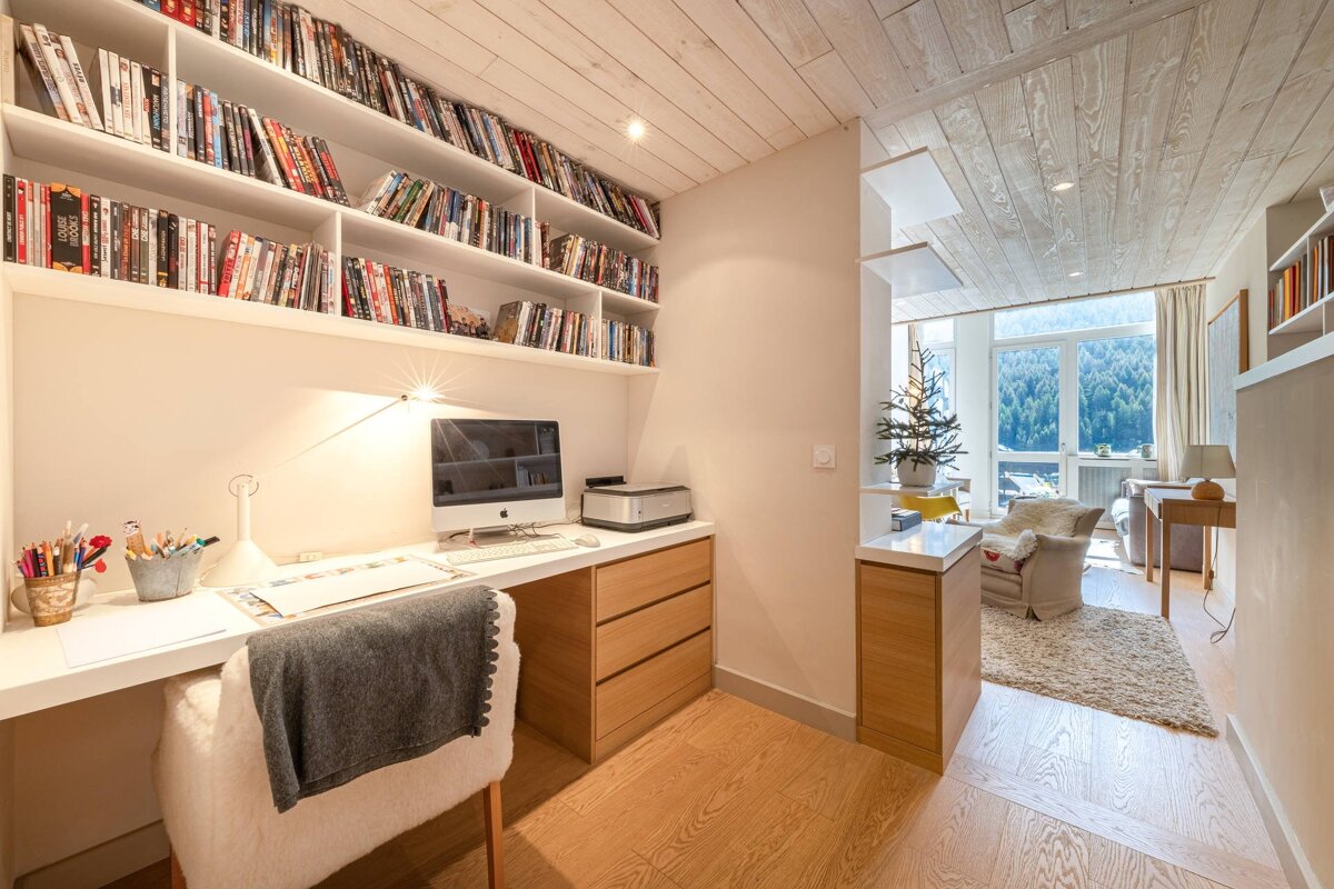 Cozy home office with wood paneling, desk, bookshelves, and a view of snowy mountains through the adjoining living area.