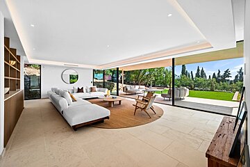 A modern living room with glass walls opening to a grassy yard, featuring sleek white furniture and neutral stone tile.