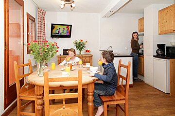 A family has breakfast in a cozy kitchen/dining area. Two kids drink juice at a wooden table, watching TV, while a woman stands in the kitchen.
