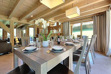 A spacious, modern dining room in a wooden chalet, featuring a large table set for dinner with an orchid centerpiece, and large windows looking out to a green landscape.
