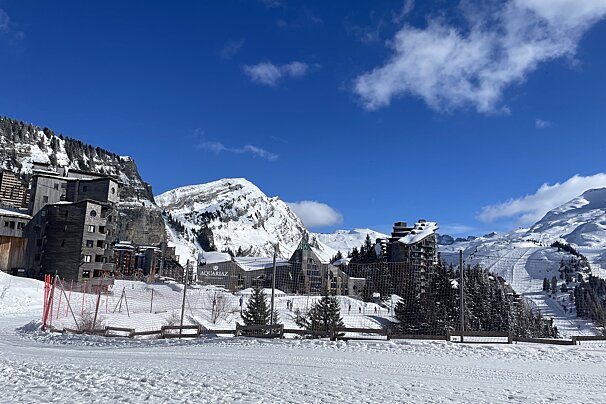 A snowy alpine ski resort with distinctive modern buildings, snow-covered mountains, and ski slopes under a bright blue sky.