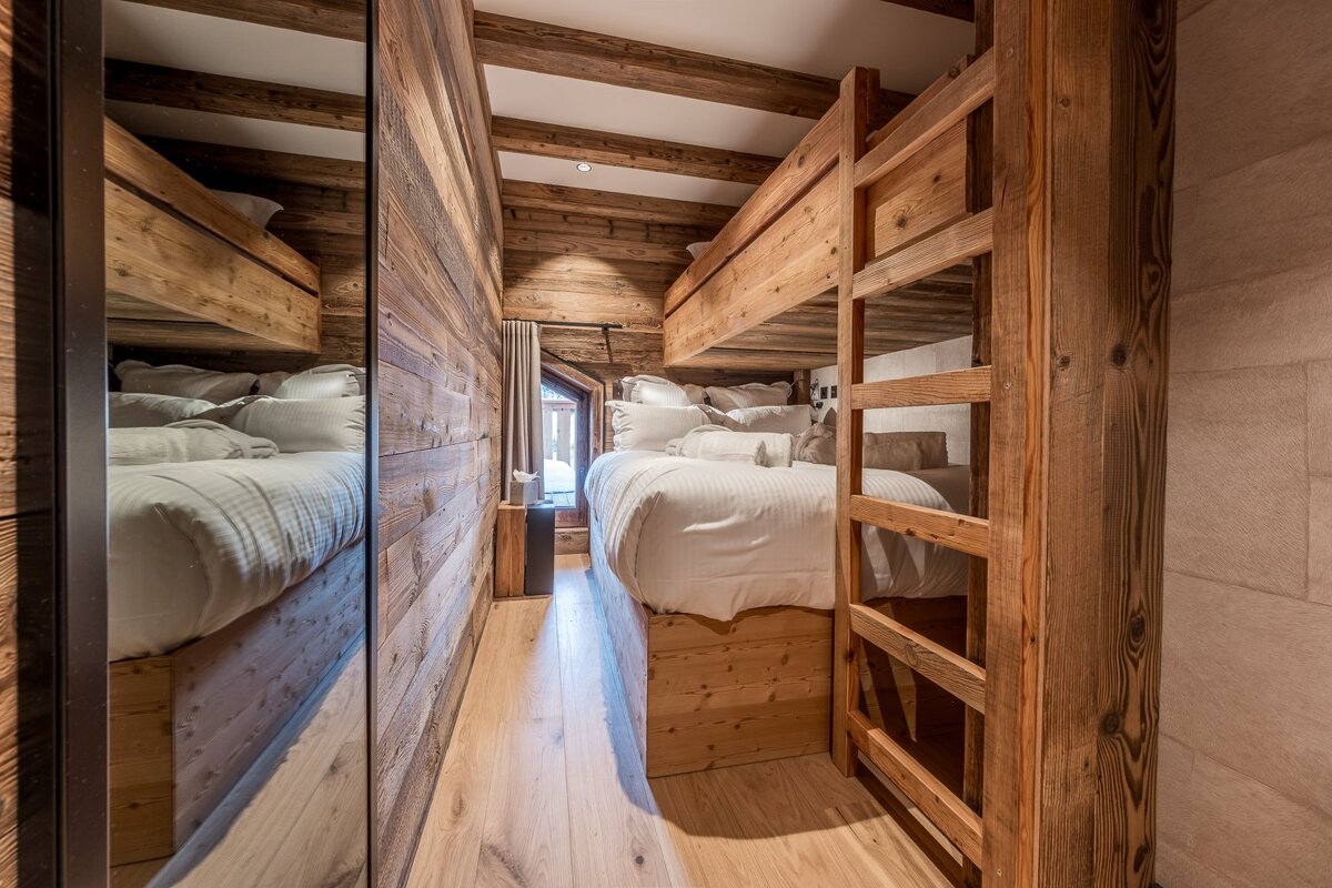A cozy, rustic wooden bedroom features built-in bunk beds with white bedding, reflected in a large mirror, with a window at the end.