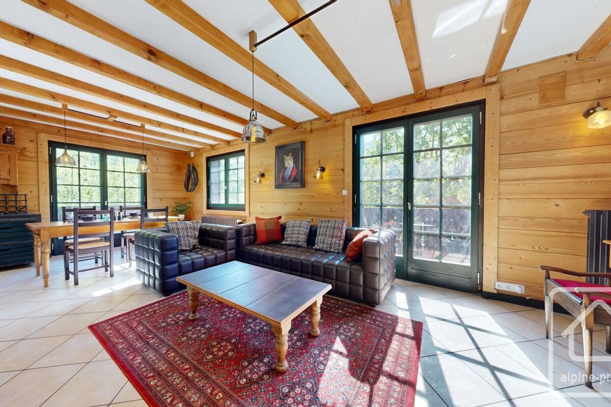 Rustic, wooden living room with a dark leather sectional sofa, a coffee table on a red patterned rug, and bright glass doors.