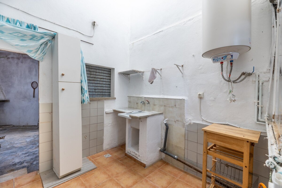 A rustic utility room features white walls, terracotta tiled floor, a double utility sink, and a large water heater. A tall cabinet and gridded window are visible.